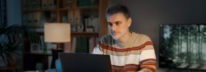 Ein Mann mit einem gestreiften Pullover sitzt an seinem Schreibtisch und schaut auf den Laptop vor ihm