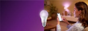 „Die Frau steuert die intelligente LED-Beleuchtung per Smartphone-App und schafft so ein warmes Licht im gemütlichen Wohnzimmer mit vernetzter Haustechnik.“